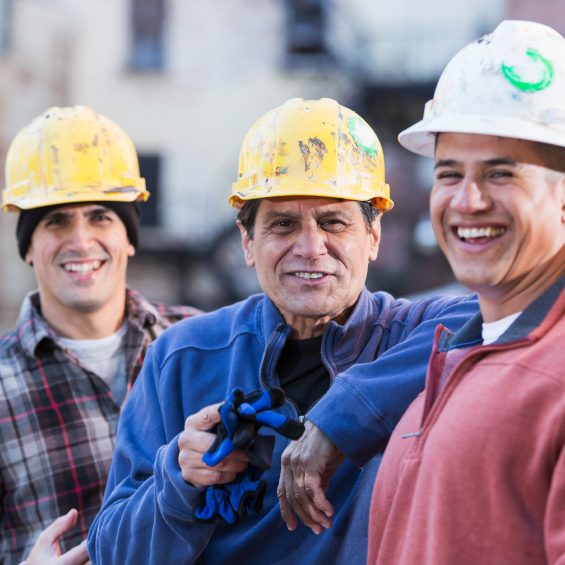 A group of three multi-ethnic men wearing white and yellow hard hats, standing in a row.  The focus is on the worker in the middle,  He is the foreman of this work crew, an Hispanic man in his 50s wearing a royal blue shirt, smiling and pointing at the camera.  They are construction workers, engineers or utility workers in the city, buildings out of focus behind them.